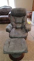 Front-facing view of gray recliner chair with matching ottoman placed on carpet. Chair has padded armrests and seat cushions with visible stitching details.