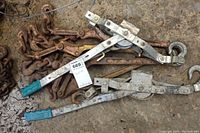 Wide view of two come along hand winches on ground with several rusty chains and load binders underneath.