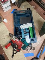 Overview of three items in the lot on floor including Drill Doctor sharpener in case, Milwaukee electric drill with cord and handle, and Makita cordless drill.