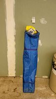 EZ Up instant shelter packed in a tall blue fabric bag standing upright against a wall.