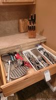 Kitchen drawer open showing wooden knife block on counter with multiple knives and drawers containing various kitchen utensils and flatware.