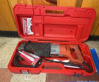 Photo showing the open red hard plastic carrying case with an orange Milwaukee Sawzall reciprocating saw inside. The saw has a power cord coiled with it.