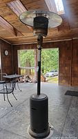 Full view of black Fire Sense propane patio heater standing on concrete porch with wooden ceiling and outdoor furniture around. Heater has round metal base, long pole, mesh heating element, and reflector top.