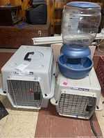 Two plastic pet carriers stacked side by side with a blue pet water bowl dispenser on top of the smaller carrier.