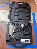 Pneumatic nail fastener inside black carrying case on floor with blue tape sections visible around.