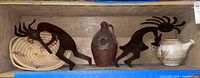 Wide view photo showing two metal Kokopelli figures on either side of pottery and woven basket on shelf.