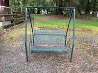 Full view of weathered green metal swing with mesh seat and backrest on a freestanding frame set outdoors on gravel.