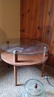 Full view of the round table showing wood lower surface and clear glass top with metal supports and three wooden legs.