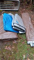 Four folded tarps in blue, brown, and silver colors on the ground outdoors, near an old vehicle part and road signs.