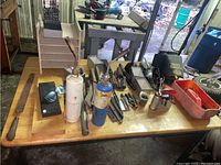 Top view of a workshop table with two oxy-fuel torches in front, multiple metal hand files, a handsaw, a yellow angle protractor, a pair of protective sunglasses, metal clamps, power tool, and a red plastic container with small parts.
