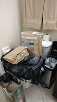 Photo showing beige heated blanket in packaging, two hanging beige towels, black duffle bag, and bathroom trash bin in corner.