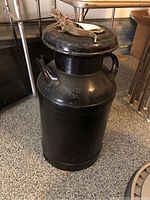 Front side view of large black antique milk can showing handles and lid with signs of wear.