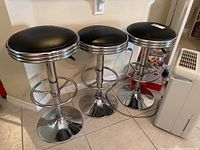 Three black and chrome bar stools shown side by side, each with round black padded seats and shiny chrome bases and footrests, adjustable height.