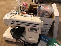 Brother XL-3030 sewing machine with foot pedal, power cord, and boxes of sewing accessories including threads and needles behind it.