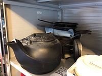 Side view of cast iron kettle with lid on shelf along with stacked skillets and other cast iron cookware