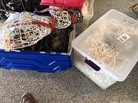 Two plastic storage bins filled with holiday decor. The left bin shows the white wire snowman partially visible inside with a red sign and red scarf. The right bin is a clear bin with a ceramic tree inside and some white string lights visible.