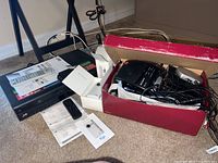 Photo showing JVC VHS player on floor with remote control and instruction manuals. Next to it is a red and white cardboard box containing a Sony electronic device and multiple tangled cables and power cords.
