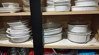 Top shelf of cabinet showing stacked white CorningWare dishes and small bowls on the left, along with oval and round baking dishes on the right.