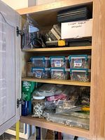 Three shelves filled with assorted screws, washers, bolts, fasteners, stapler and staples organized in containers and boxes inside a wood cabinet.