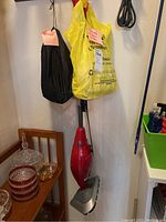 Full view of red Dirt Devil steam mop hanging on wall with attached power cord and yellow plastic bag with accessories.