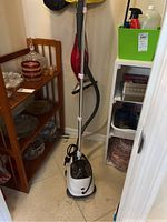 Full view of the clothes steamer standing upright on the floor next to shelves with household items.