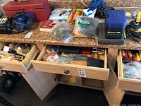 View of three drawers and two cabinets opened showing numerous tools including gloves, bags of hardware, metal rivet drivers, a blue plastic case, red handled tools, and various hand tools and hardware scattered inside drawers and on countertop.