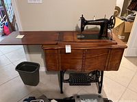 Full view of vintage sewing machine mounted in wooden cabinet with tabletop extension and black plastic trash bin on floor.