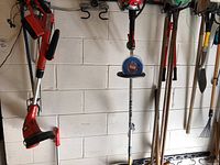 Red and black cordless weed wacker hung on wall with battery pack and handle visible with other long handled tools like rakes and shovels.