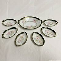 Top-down view of the antique Bavarian berry dessert set arranged with one large handled serving dish in the center and six smaller oval dishes surrounding it. Pieces have floral patterns with green and gold decorative edges.