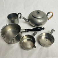 Overview of the pewter teapot with bamboo handle, measuring cup, saucepan with black handle, and two small measuring pitchers with spouts and handles arranged on a white surface.