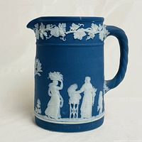 Side view of Wedgwood blue jasperware pitcher featuring white classical figures in relief, including a woman holding a basket and another standing near a small fire.