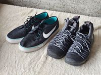 Two pairs of sneakers together on a white textured surface, Nike canvas low-tops black with white swoosh and teal interior compared to gray Under Armour knit high-tops with thick laces.