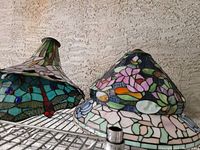 Two Tiffany style stained glass lampshades on a shelf against a textured wall. Left lampshade has dragonfly motif in green and blue glass with red accents. Right lampshade has floral pattern in pink, blue, orange, and green glass pieces.
