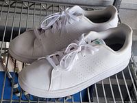 Side angled view of white Adidas sneakers with green branding placed on shelf