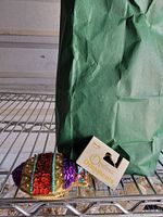 Photo showing one example ornament: a multicolored sequined turtle with original Ovente Ornaments tag attached, resting on a metal wire shelf next to a green bag containing other ornaments.