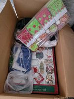 Photo shows a cardboard box full of assorted Christmas decorations including a box of 40 LED clear string lights, several ornaments in green and red packaging, and multiple Christmas figurines.