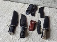 Overview of five knives with corresponding sheaths and one brown leather pouch, laid out on white fabric