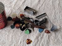 Photo showing various guitar picks spread out with a wooden string winder and metal capo-like accessory on a soft fabric background.