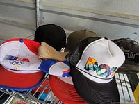 Seven assorted hats on a metal shelf, various colors and logos visible including USA Olympic McDonald's and DON mesh caps.