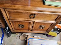 Wide shot showing the lower drawers and brass ring pull hardware of one nightstand, on a concrete floor with some other stored items visible around.