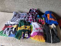 Lot of assorted sweatshirts including Nintendo 64, Mickey Mouse, NBA, colorful stripes, and plaid shirt, laid out on beige surface.
