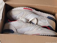 Side view of Reebok sneakers in original box showing beige and white mesh and suede panels, red accents, and rubber sole with wave tread.