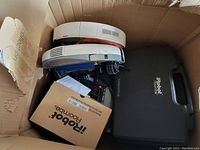 Top-down view of two white Roomba units stacked inside a cardboard box with a black hard carrying case branded iRobot Roomba.