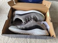 Pair of gray Skechers sneakers placed side-by-side inside original brown cardboard box with lid open, showing white sole and light wear on shoes.