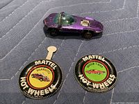 Top-side view of purple Hot Wheels Silhouette car with visible wear, placed above two Mattel Hot Wheels tin buttons labeled Silhouette and Custom T-Bird.