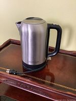 Front and side view of the stainless steel electric tea kettle placed on a wooden table with power cord attached.