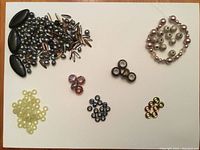 Assortment of jewellery beads laid out on a white surface showing a variety of bead shapes, sizes and colors including black oval beads, pearl-like beads, and small round beads in yellow, pink, black and amber hues.