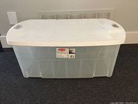 Clear Jumbo Rubbermaid plastic tote with white lid sitting on a dark carpeted floor.