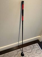 Full length view of the right handed golf club with black shaft and red-black grip showing the entire club including clubhead on floor
