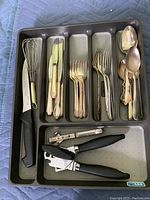 Top-down photo of a gray plastic cutlery tray containing a black handled knife, metal whisk with cream handle, three knives with cream handles, six forks, three decorative spoons, and two black handled can openers